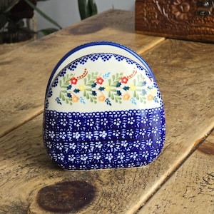 Puede incluir: Un servilletero de cerámica con un diseño floral azul y blanco. La sección superior presenta un fondo blanco con flores coloridas y hojas verdes. La sección inferior tiene un fondo azul con un patrón floral blanco.