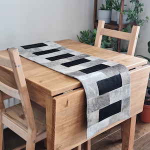 May include: A black and white checkered table runner made of cowhide leather. The runner is on a wooden table with a wooden chair.