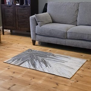 May include: Rectangular area rug with a geometric pattern in shades of gray and white. The rug is placed on a wooden floor in front of a gray sofa. The pattern radiates from a central point.