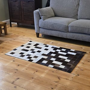 May include: A rectangular rug with a geometric pattern of black, white, and brown squares. The rug is made of a natural material and is placed on a wooden floor. The rug is in front of a gray sofa.
