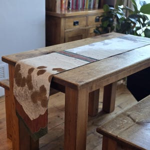 May include: A wooden table with a cowhide-patterned table runner. The runner has brown and white spots, with a red and green border. The table is made of light brown wood and has a matching bench. A wooden cabinet and a plant are in the background.