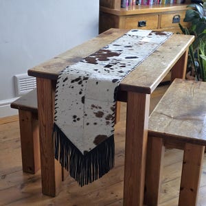 Könnte beinhalten: Ein Tischläufer aus Kuhfell mit einem weiß-braunen Fleckenmuster. Der Läufer hat am Ende eine schwarze Fransen und liegt auf einem Holztisch. Der Tisch ist von Holzbänken und einem Holzboden umgeben.