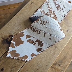 May include: Two cowhide table runners with brown and white patterns. The runners are stitched together with brown thread and have a leather-like trim. The runners are on a wooden table.