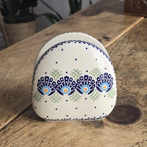 Puede incluir: Servilletero de cerámica color crema con un patrón decorativo. El diseño presenta motivos florales azules y naranjas y pequeños puntos azules. El soporte está sobre una superficie de madera.