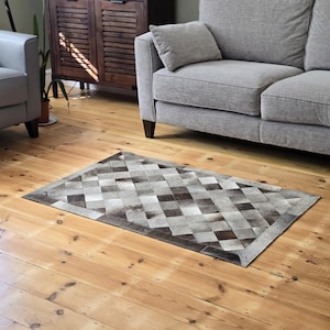 May include: A grey and brown cowhide rug with a diamond pattern. The rug is laid out on a wooden floor in a living room setting.