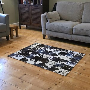 Peut inclure: Un tapis rectangulaire en peau de vache avec un motif patchwork. Le tapis présente un motif de pièces noires et blanches, créant un aspect quadrillé. Le tapis est posé sur un plancher en bois.