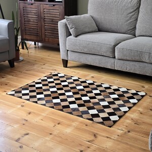 Op de afbeelding: Een bruin, zwart en wit koeienhuidkleed met een ruitpatroon. Het kleed ligt op een houten vloer voor een grijze bank.