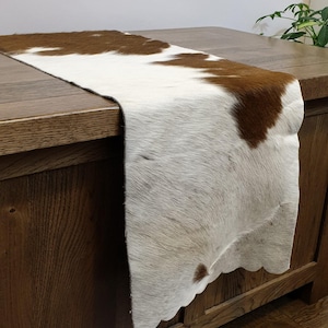 May include: A cowhide table runner in natural brown and white tones. The runner is laid across a wooden surface, highlighting the hide's texture and colour variations. The edges are gently scalloped.