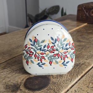 May include: A white ceramic napkin holder with a blue rim and a floral design featuring red berries, blue leaves, and yellow accents. The holder is on a wooden surface.