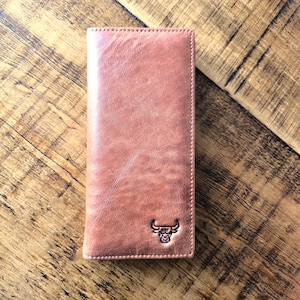 May include: A brown leather wallet with a bull head logo. The wallet is rectangular and has a vertical design. The leather has a textured appearance. The wallet is on a wooden surface.