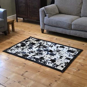 May include: Rectangular cowhide rug with a black border. The rug features a patchwork design of black and white cowhide squares, creating a checkered pattern. The rug is placed on a wooden floor.