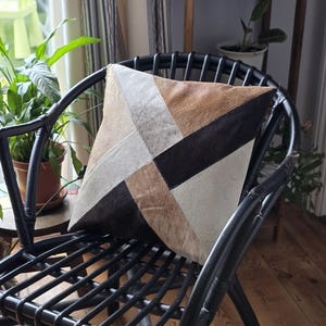 Può includere: Una sedia in vimini nera con un cuscino in pelle patchwork marrone, nero e bianco. Il cuscino ha un motivo geometrico.