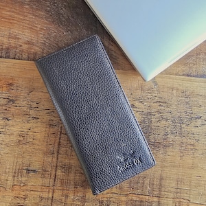 May include: A dark brown leather wallet with a textured finish. The rectangular wallet has the words "CROSS OX" embossed on the lower right corner. A laptop is visible in the background.