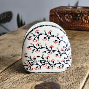 Peut inclure: Un porte-serviettes en céramique avec un motif floral. Le support est blanc avec une bordure noire et présente un motif de fleurs rouges et de feuilles vertes sur des tiges noires. Le support est sur une surface en bois.