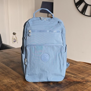 May include: A light blue backpack with multiple zippered compartments and a top handle. The backpack features a front pocket, side pockets, and a logo patch. The bag is made of a durable, water-resistant material.