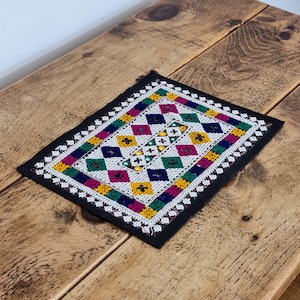 May include: A colourful embroidered fabric square with a black border. The fabric is decorated with a geometric pattern of diamonds in shades of pink, green, yellow, blue, and white. The fabric is embellished with beads and sequins.