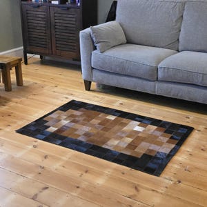 May include: Rectangular area rug with a patchwork design. The rug features squares of brown, tan, and white, framed by a black border. The rug is placed on a wooden floor, near a gray sofa.