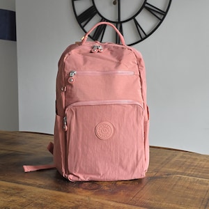 May include: A dusty rose backpack with multiple zippered compartments and a top handle. The backpack features a front pocket with a circular logo and side pockets. The bag is made of a durable, water-resistant material and is designed for everyday use.