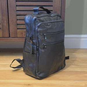 May include: A dark gray leather backpack with multiple zippered compartments and a top handle. The backpack has adjustable shoulder straps and a small pocket on the side. The leather has a textured appearance.