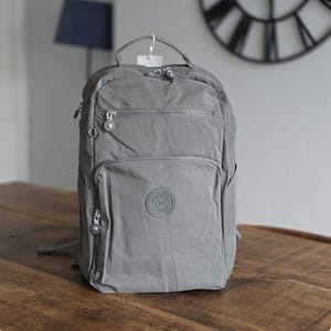 May include: A grey backpack with several zipped compartments and a top handle. The backpack features a front pocket and side pockets, and a small logo patch. The bag is made of a smooth, durable material, suitable for daily use.