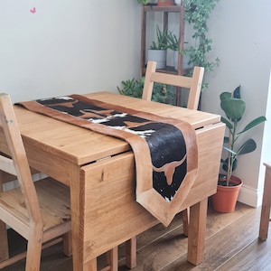 May include: A cowhide table runner with a black and white pattern and brown leather trim. The runner is displayed on a light wood table, showcasing its rustic design. The table is surrounded by wooden chairs.