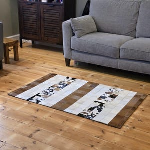 May include: Rectangular cowhide rug with a striped pattern. The rug features alternating stripes of brown, white, and sections with black and brown speckled patterns. The rug is placed on a wooden floor, with a gray sofa and wooden cabinet in the background.
