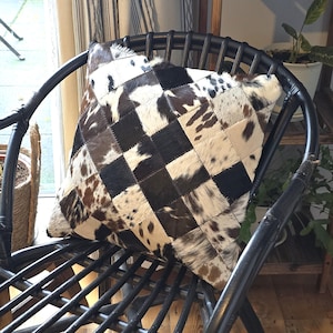 May include: A black and white patchwork cowhide cushion with a diamond pattern. The cushion is sitting on a black wicker chair.