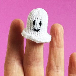 May include: A white knitted ghost finger puppet with a smiling face. The ghost has a simple design with a rounded body and a ruffled bottom edge. The puppet is on a pink background.