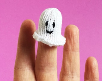 May include: A white knitted ghost finger puppet with a smiling face. The ghost has a simple design with a rounded body and a ruffled bottom edge. The puppet is on a pink background.