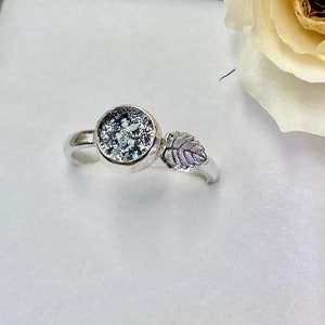 May include: A silver ring with a round, blue and white stone and a leaf design. The ring is in a white box with a white rose.