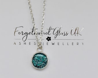 May include: A silver necklace with a round pendant filled with teal glitter. The pendant is set in a silver bezel and hangs from a delicate silver chain. The necklace is displayed on a white box with the text "Forgetmenot Glass UK" and "Ashes Jewellery".