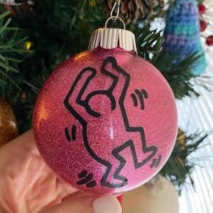 May include: Pink Christmas ornament with a black and white stick figure design. The ornament is covered in glitter.