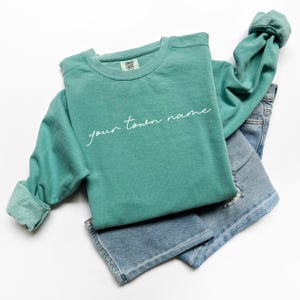 May include: A teal green sweatshirt with the text "your town name" in white script on the front. The sweatshirt is folded and laid on top of a pair of blue jeans.