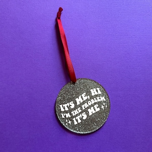 Könnte beinhalten: Silberner Glitzer-Weihnachtsbaumschmuck mit rotem Band. Der Schmuck hat den Text "It's Me, Hi I'm the Problem It's Me" in weiß gedruckt.