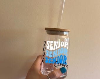 May include: A clear glass tumbler with a bamboo lid and a clear straw. The tumbler has the words "Senior 2026" in white and blue, with the name "Sarah" in black. A ring is on the finger holding the tumbler.