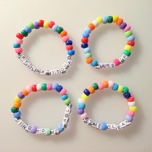 May include: Four circular bracelets made of multicolored plastic pony beads. Each bracelet features white square letter beads spelling out phrases: WFYILWFRITDUAD, WAKE UP, SING TO ME PAOLO, and COME CLEAN. The beads are arranged in a repeating color pattern.