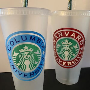 以下が含まれることがあります： ふた付きストロー付きの透明なプラスチックカップが2つ。カップには、女性の顔と星がついた緑色のスターバックスロゴがあります。カップのテキストは、青色の「Columbia University」と赤色の「Harvard University」です。