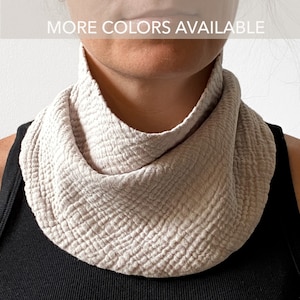 May include: A beige neck scarf featuring a textured, crinkled fabric finish that drapes around the neck. The scarf is worn over a black tank top. The image includes the text MORE COLORS AVAILABLE at the top, indicating additional options.