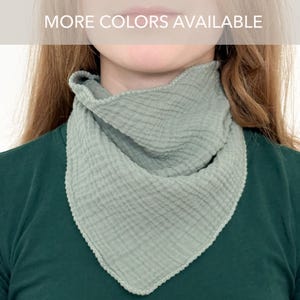 May include: A sage green triangular bandana scarf made of textured muslin fabric with a crinkled appearance and a finished stitched edge. The scarf is worn around the neck over a dark green shirt. The text MORE COLORS AVAILABLE is visible.