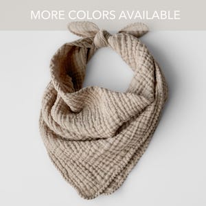 May include: A beige muslin bandana bib with a knotted top. The bib is made of a soft, textured fabric and is designed to catch drool. The text "MORE COLORS AVAILABLE" is at the top.