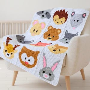 May include: A white baby blanket with a repeating pattern of animal faces, including a bear, fox, and rabbit. The blanket is draped over a cream-colored armchair in a nursery setting. The blanket is made of a soft, textured material.