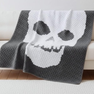 Könnte beinhalten: Eine grau-weiße Häkeldecke mit einem Totenkopf-Design, die über einem weißen Sofa drapiert ist. Der Totenkopf ist weiß, mit grauen Augenhöhlen und Nase. Der Hintergrund der Decke ist grau. Eine Halloween-Fledermausgirlande hängt im Hintergrund.
