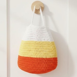 Puede incluir: Un bolso tote de ganchillo hecho a mano con una combinación de colores de maíz dulce: blanco, amarillo y naranja. El bolso está colgado de una clavija de madera en una pared blanca. El bolso tiene un fondo redondeado y asas largas.
