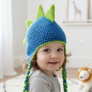 May include: A blue and green crochet dinosaur hat with ear flaps. The hat features a blue body with green spikes and trim. The ear flaps are braided with blue and green yarn. A child is wearing the hat.