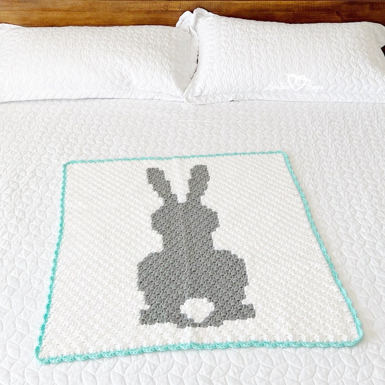 May include: A square crochet blanket featuring a white background with a central grey silhouette of a rabbit. The blanket has a textured stitch pattern and is finished with a bright turquoise border around all four edges of the square fabric piece.