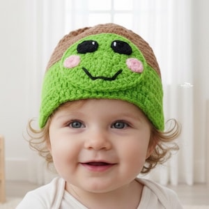 Puede incluir: Un gorro de bebé de ganchillo verde y marrón con un diseño de cara de rana. El gorro presenta ojos negros, mejillas rosas y una boca sonriente. El gorro lo lleva un bebé con ojos azules y pelo rubio rizado.