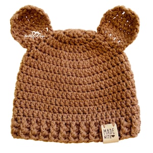 May include: A brown crochet hat with bear ears. The handmade hat has a tag that reads "MADE WITH LOVE". The hat is designed for babies and toddlers. The hat is made of a soft, textured yarn.