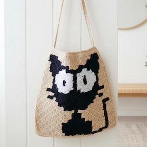 May include: A beige crochet tote bag featuring a black cat design with large white eyes. The bag has long beige straps and is hanging against a white wall. The cat design is a prominent feature.