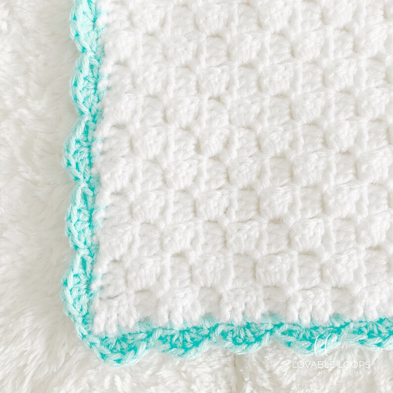 May include: A white crocheted blanket featuring a textured square stitch pattern and a scalloped border in a bright turquoise yarn. The corner of the blanket is clearly visible, showcasing the intricate hand-stitched detail and the colour contrast.