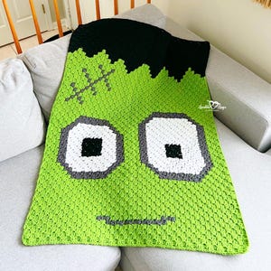 May include: A handmade, green crochet blanket featuring a Frankenstein monster design. The blanket has black hair, gray stitches, and large eyes with white, gray, and black details. The blanket is displayed on a gray couch.